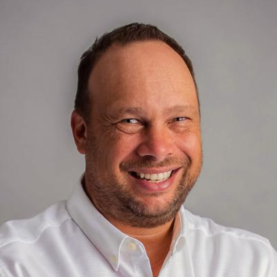 A man with short, brown hair and a light beard is smiling while wearing a white button-up shirt. The background is a plain gray.