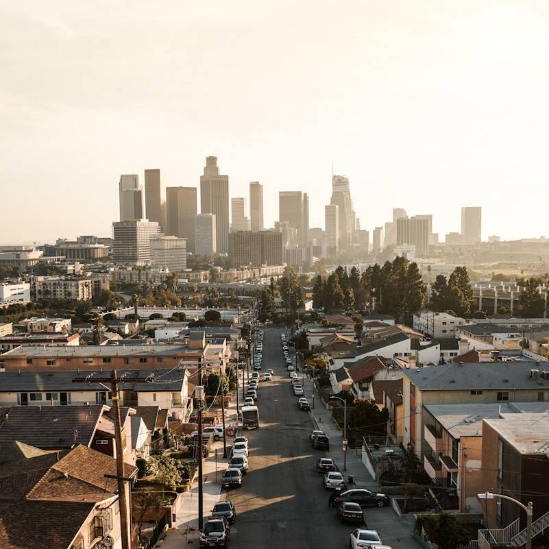 Aerial shot of a city street lined with parked cars and houses, leading toward the downtown area with tall skyscrapers in the distance. The scene is bathed in a warm, golden light, suggesting either sunrise or sunset. Trees and small buildings are visible in the foreground.