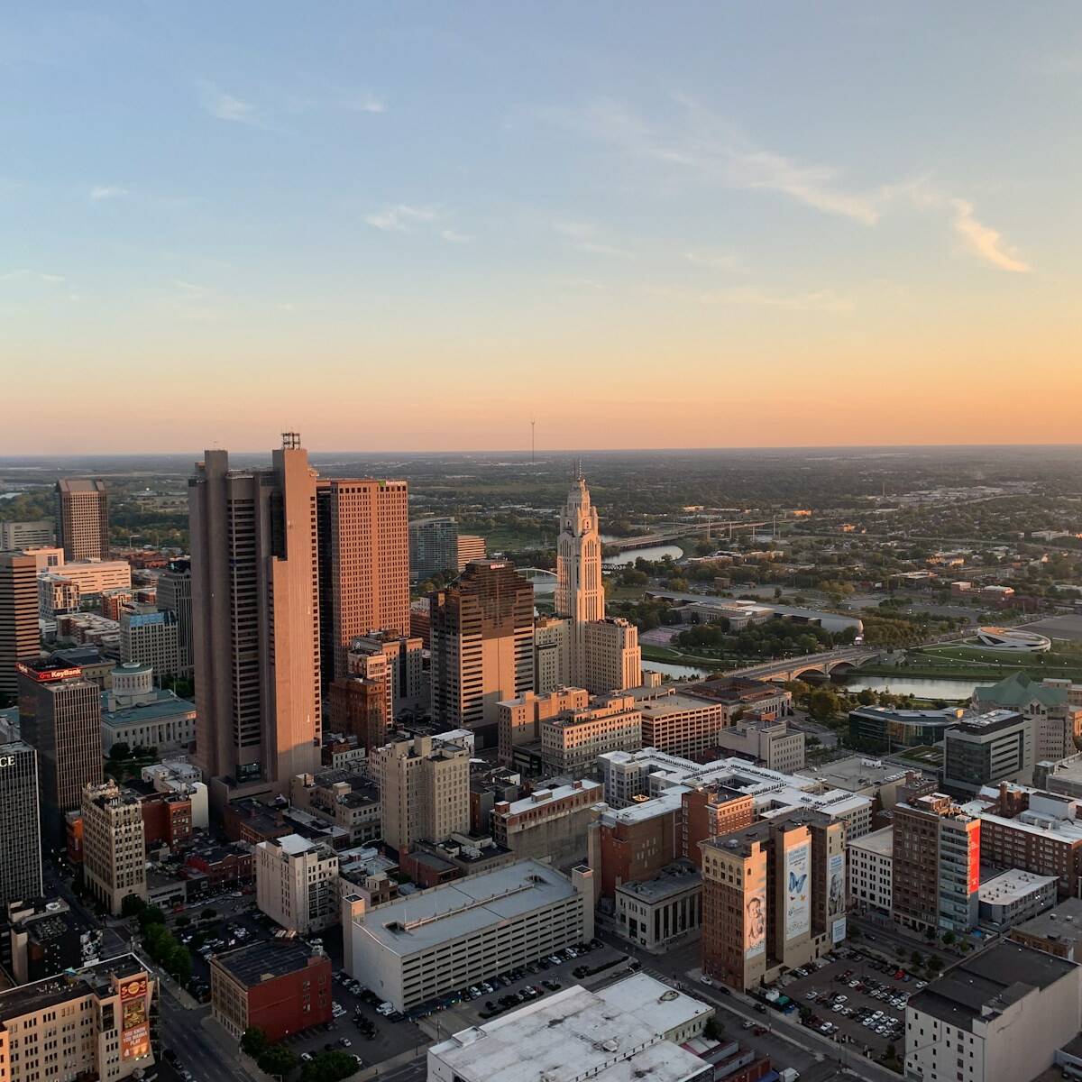 Aerial view of a cityscape at sunset. Tall buildings and skyscrapers are clustered in the foreground, tapering off to smaller structures and greenery in the distance. The sky features soft clouds and a warm, orange hue from the setting sun.