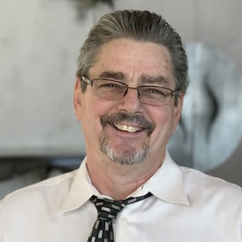 A man with short, graying hair, glasses, and a mustache is smiling. He is wearing a white dress shirt and a patterned tie, which is loosely knotted. The background appears to be indoors and somewhat out of focus.