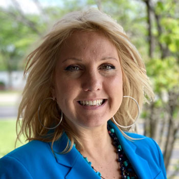 A woman with blonde hair smiling at the camera. She is wearing a blue blazer, large hoop earrings, and a beaded necklace. The background is blurred with greenery.