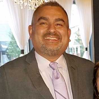 A man in a gray suit with a light purple tie is smiling at the camera. He stands in front of a window with white curtains. The background appears to be a bright, elegant indoor setting.