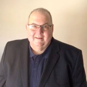 A person with short hair and glasses is standing against a plain beige background. They are wearing a dark blazer over a dark collared shirt and are smiling at the camera.