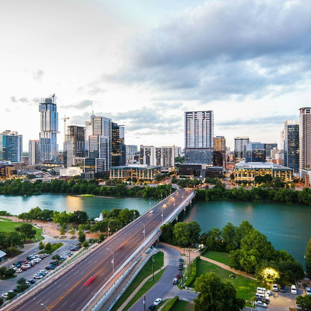 Aerial view of a cityscape featuring a river bordered by green parkland and a bridge with traffic. Tall buildings and skyscrapers fill the background under a cloudy sky, reflecting the urban landscape. Trees and a parking area are visible in the foreground.