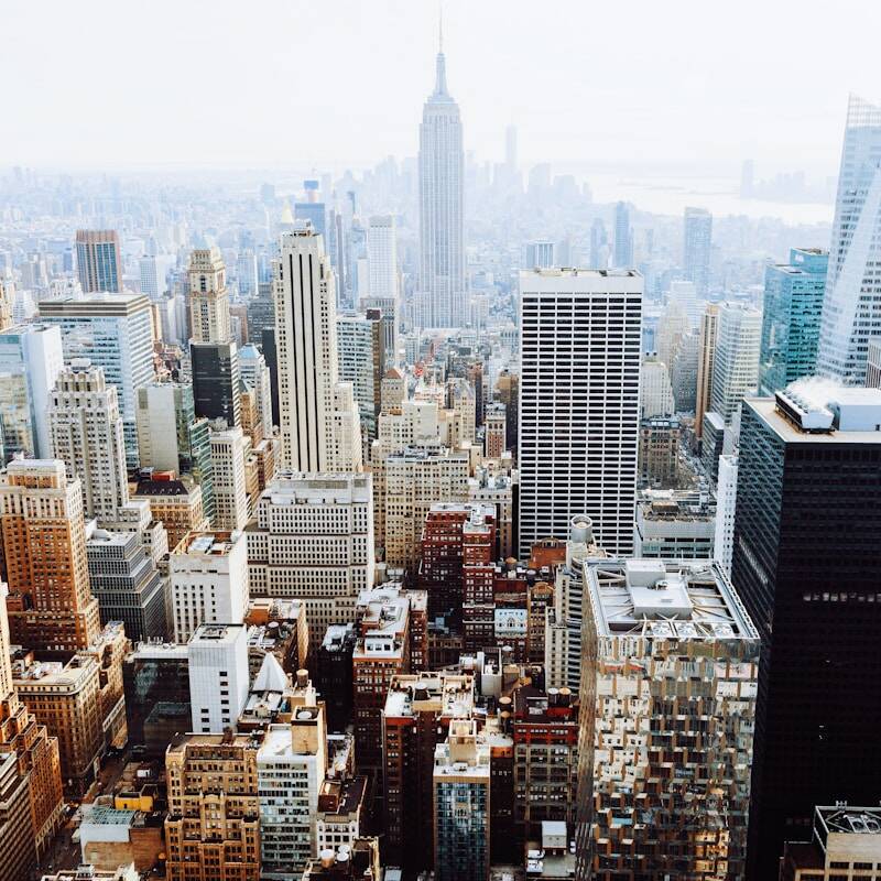 Aerial view of a city's skyline dominated by numerous skyscrapers and high-rise buildings. The tallest building, centrally located, stands prominently among the rest. The image captures the urban density and architectural diversity during daytime.