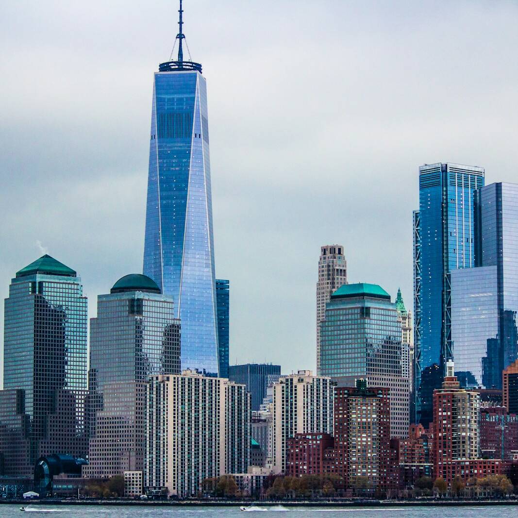 A cityscape featuring New York City's Lower Manhattan skyline with prominent skyscrapers, including the tall, blue-tinted One World Trade Center, and other surrounding buildings under a cloudy sky. The Hudson River is visible at the bottom edge of the image.