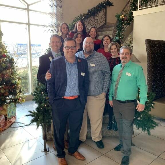 A group of eleven adults, some smiling and some neutral, pose together in front of a staircase decorated with holiday-themed garlands at the 2018 holiday luncheon. There is a Christmas tree to the left and a large window in the background letting in natural light.