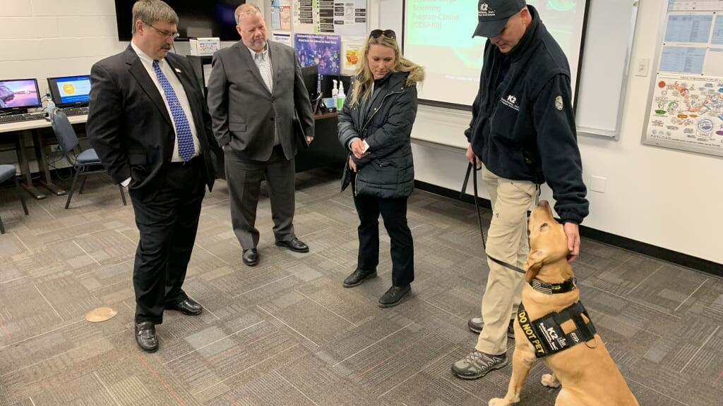 Four individuals in business attire stand in a room, three of them observing a handler in uniform with a yellow labrador retriever wearing a working dog vest. The room has office equipment and a large screen displaying a presentation.