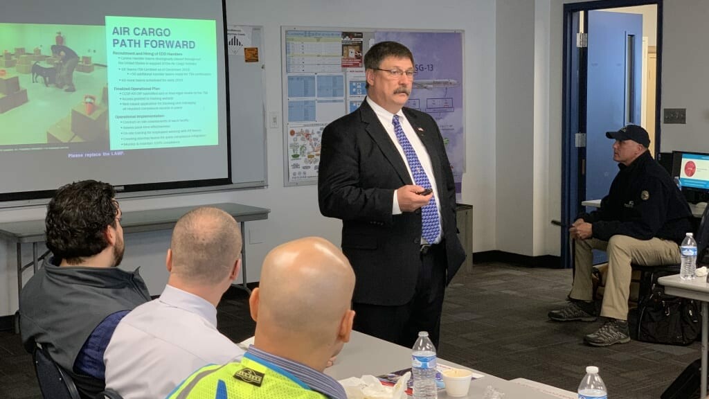 A man in a suit is giving a presentation in a classroom-like setting. The slide behind him reads 