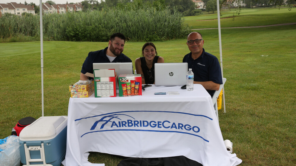 Three people sit under a canopy at an outdoor event, working on laptops behind a table covered with an 