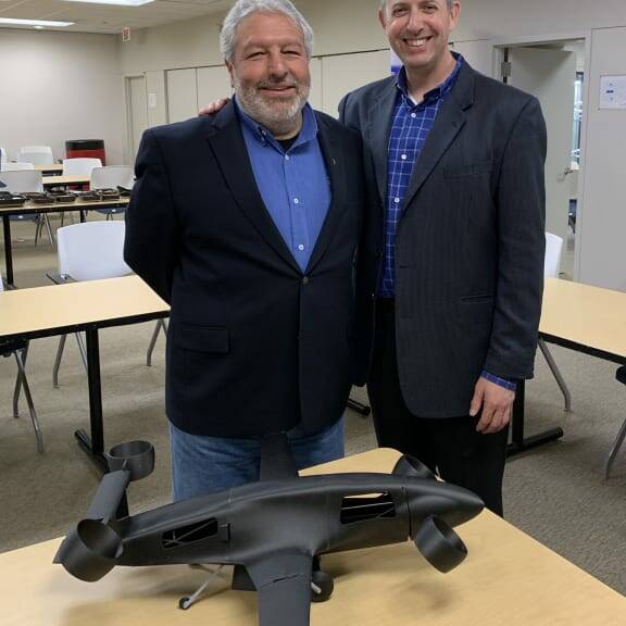 Two men are standing indoors, smiling at the camera during a 2019 luncheon. One man is wearing a navy blazer and blue shirt, while the other is in a dark suit with a blue checked shirt. In front of them on a table is a small, black model of an aircraft with vertical landing capabilities.