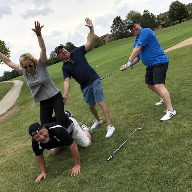 During a 2018 golf outing, four people on the course engage in playful antics. One man is on all fours, another seated on his back, while a woman balances on one foot with her arms raised. Meanwhile, a fourth man playfully pulls a golf club from the seated man's waist. #IACAC