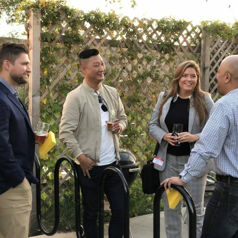 Four people are standing outside and engaged in conversation. Three men and one woman seem to be enjoying drinks together at what appears to be a September Oktoberfest event. They are casually dressed and appear to be at a social gathering or work event, with greenery and a wooden lattice fence in the background.