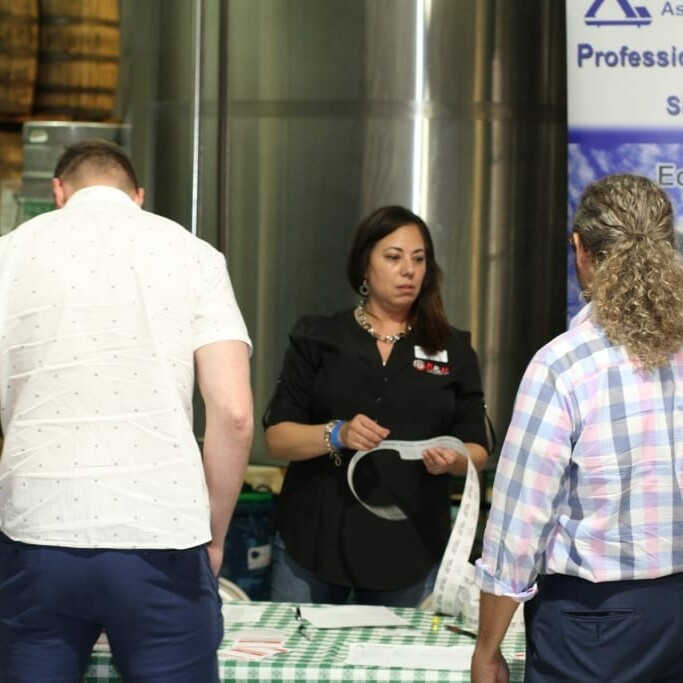 A woman stands behind a table with a green-checkered tablecloth, engaging with two men in front of her. Behind the woman, an IACAC banner promotes professional networking, education, and community. The lively atmosphere feels reminiscent of Oktoberfest 2018.