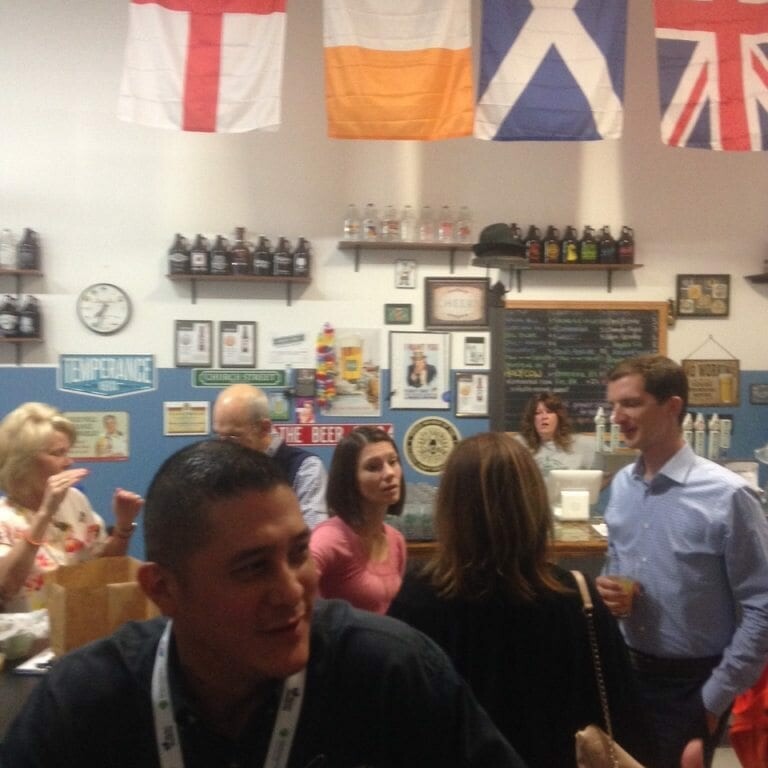 A group of people are socializing inside a room with various flags hanging on the wall, including those of England, Ireland, Scotland, and the United Kingdom. It's November 2022, and there are framed pictures and a menu board on the wall behind them.