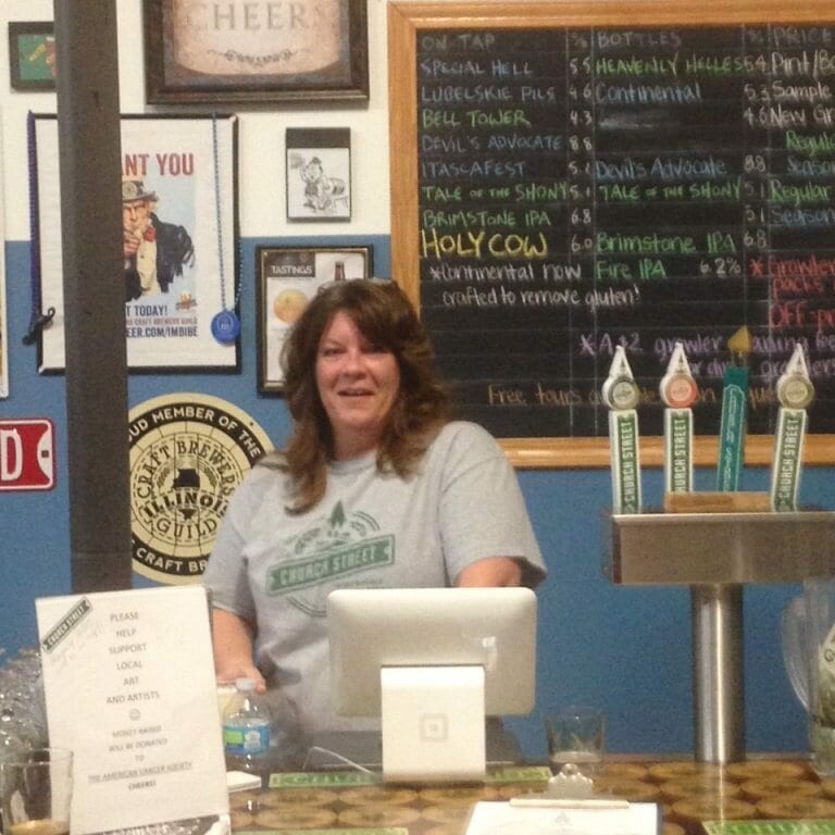 A woman with shoulder-length hair wearing a gray T-shirt stands behind a bar counter equipped with a point-of-sale system. Behind her is a chalkboard menu listing various beer options from the 2022 Oktoberfest and taps with labeled handles. The bar has various signs and decorations on the walls.