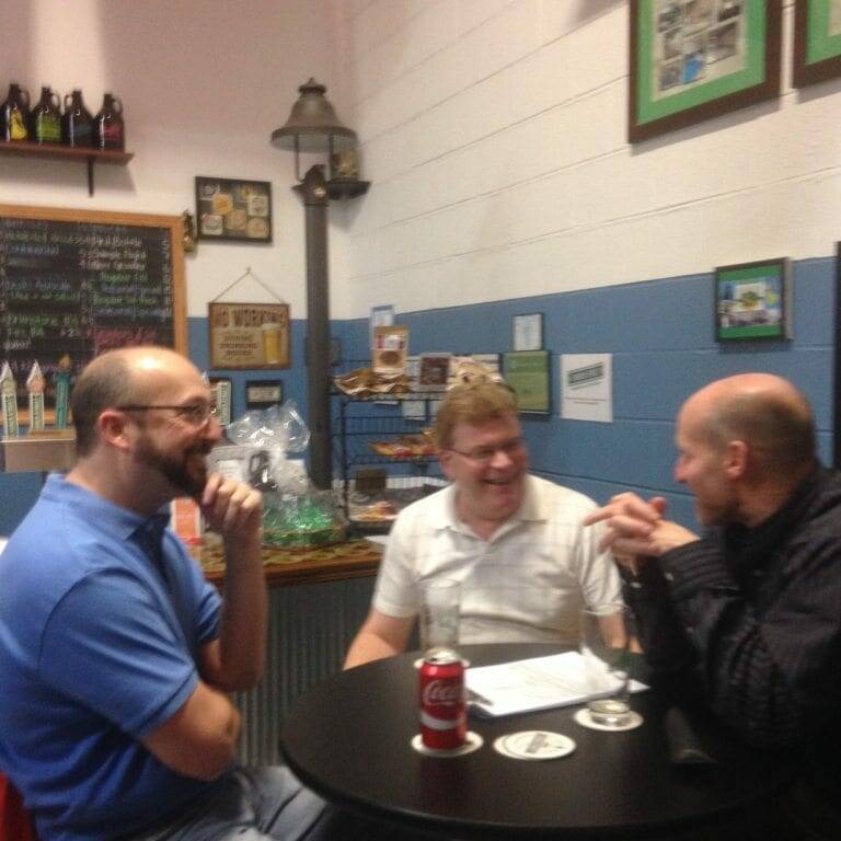 Three men sit around a round table in a casual indoor setting, engaged in conversation. One holds a can of soda, and the table has drinks and papers on it. The background features a wall with framed pictures, a Union Jack flag, and other decor items, capturing the essence of November 2022.