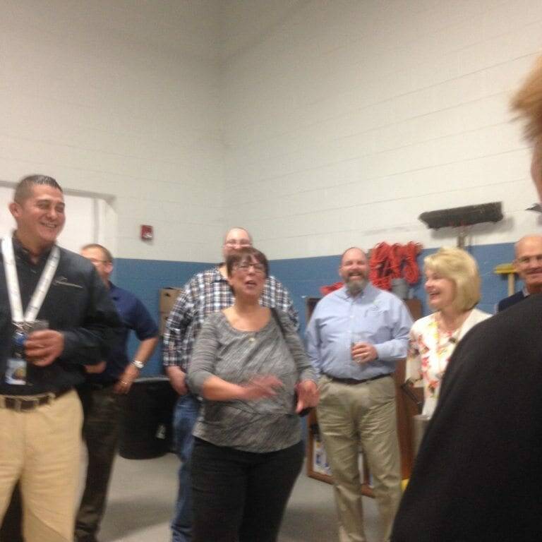 A group of people is gathered in a room, many smiling and engaging in conversation. The woman in the center appears to be speaking, while others listen attentively. The setting seems informal, with some wearing badges. Industrial elements are visible in the background, hinting at a unique 2022 Oktoberfest celebration.