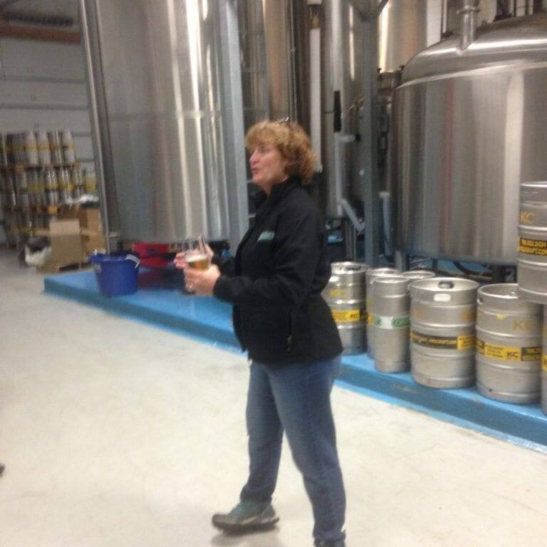 A person with curly hair, wearing a black jacket, jeans, and teal shoes, is standing in a brewery during November 2022. They hold a small bottle while speaking. Behind them are large silver brewing tanks and several beer kegs stacked and arranged throughout the area.