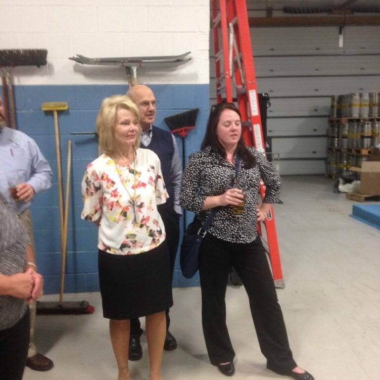 A group of people standing in a workshop environment with a ladder and cleaning tools against the wall in the background. A woman in a floral blouse and black skirt, reminiscent of Oktoberfest attire, stands next to a man in a suit. Another woman in a black outfit holds a glass.