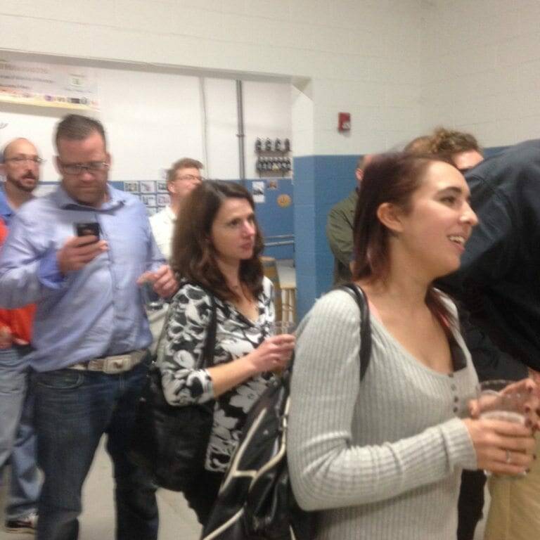 A group of people stands indoors, some holding drinks. One person is looking at a phone while others appear to be engaged in conversation. The casual setting features both men and women, with a white and blue wall and some posters or signs in the background—a perfect snapshot of an Oktoberfest gathering in November 2022.