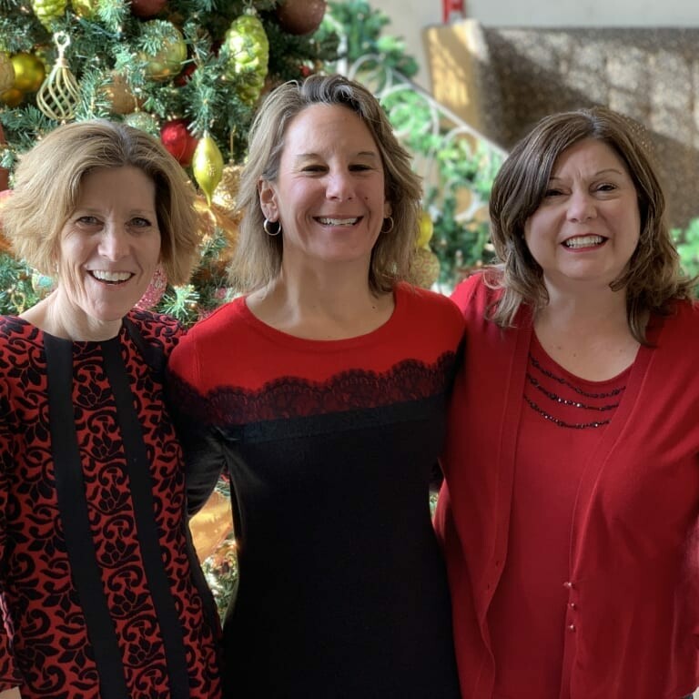 Three women smiling and standing in front of a decorated Christmas tree during a 2018 holiday luncheon. They are dressed in red and black festive outfits, and the background features holiday decorations and warm lighting.