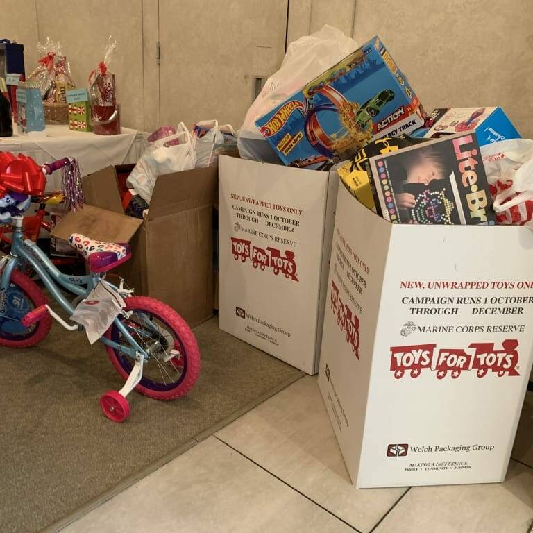 A collection of toys is displayed for a 2018 holiday donation drive. A decorated pink and blue bicycle stands beside two large donation bins filled with new, unwrapped toys. The bins are labeled "Toys for Tots," with more toys and gifts visible in the background, creating a festive luncheon-like atmosphere.