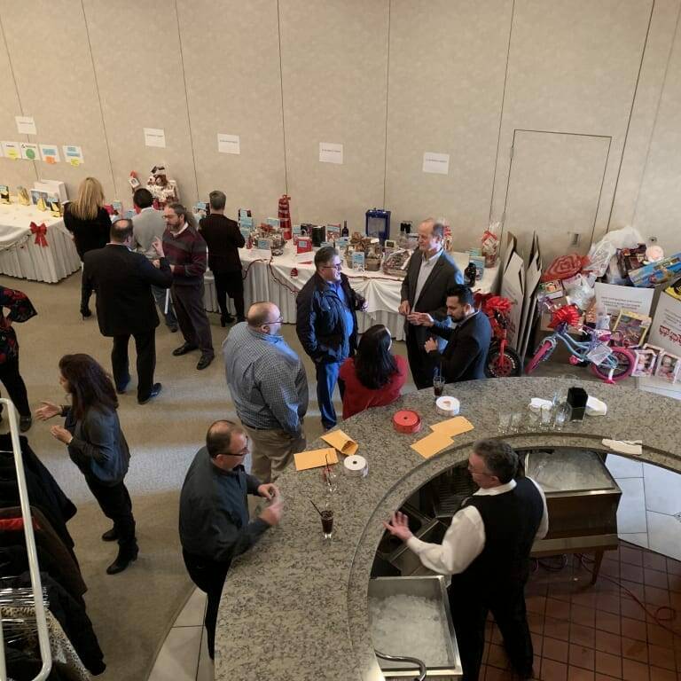 A group of people talking and browsing near tables laden with various items in a banquet hall during the 2018 holiday luncheon. Gift baskets, wrapped presents, and other raffle items are displayed on the tables, while a bartender serves drinks at a bar in the foreground.