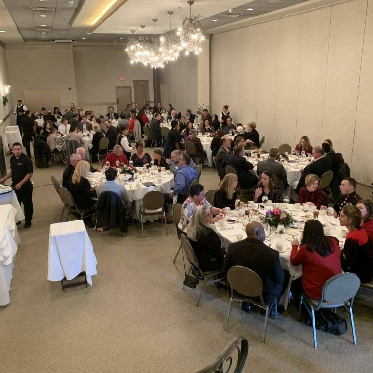 A large banquet hall filled with people sitting at round tables, engaged in conversation and dining. Servers attend to the guests as tables decorated with flowers and tableware capture the eye. The atmosphere is lively and bustling, reminiscent of a grand holiday luncheon from 2018.