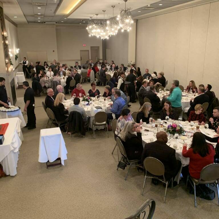 A large banquet hall filled with people seated at round tables covered with white tablecloths during a holiday luncheon. Guests are dining and engaging in conversation. Servers dressed in black are attending to the tables. Decor includes elegant lighting and floral centerpieces, reminiscent of 2018.