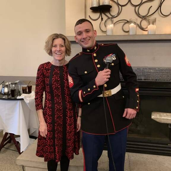 A woman in a red and black patterned dress stands next to a man in a formal military uniform at the 2018 Holiday Luncheon. They are both smiling, with the man holding a microphone. They are indoors, standing in front of a fireplace adorned with decorative ironwork and candles above it.