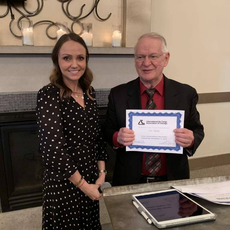 Two people are standing indoors. The person on the right, dressed in a black suit with a red shirt and tie, is holding a certificate from the 2018 holiday luncheon. The person on the left, wearing a black dress with white polka dots, is smiling at the camera. A mantel with candles is in the background.