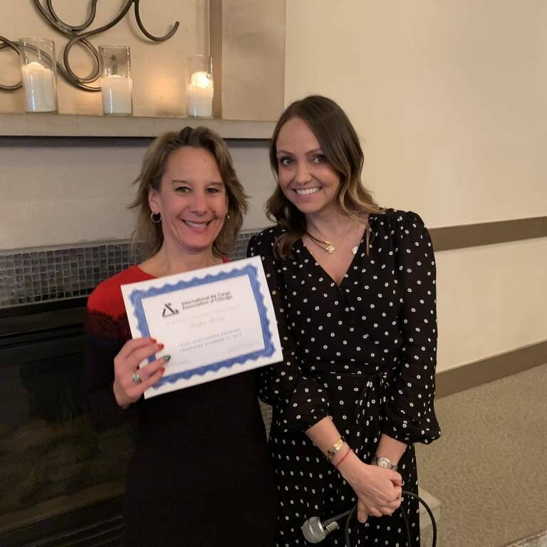 Two women standing indoors during a holiday luncheon. One woman is holding a certificate from 2018 with a blue border, smiling, and wearing a red-and-black dress. The other woman, in a black dress with white polka dots, is standing beside her, also smiling. A fireplace with candles is in the background.