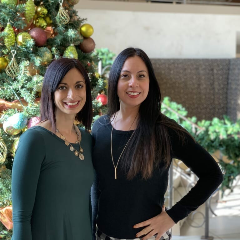 Two women are standing and smiling in front of a beautifully decorated Christmas tree, capturing the joyous 2018 holiday spirit. The woman on the left is wearing a dark green dress with a statement necklace, while the woman on the right sports a black top with a long necklace and plaid pants.