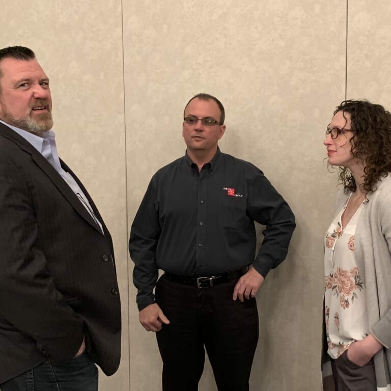 Three people are standing and conversing in what appears to be a professional setting. The man on the left is wearing a dark suit, the man in the middle is in a dark shirt with a logo, and the woman on the right is in a light cardigan and floral blouse, all possibly discussing plans for an upcoming holiday luncheon.