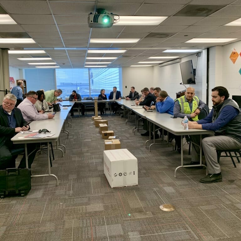 A group of people sitting around a large rectangular table in a conference room during the 2019 February Luncheon by K2 Solutions. Some individuals are working on laptops while others are engaged in conversation. The room has large windows and a ceiling-mounted projector. Boxes and packages are on the floor.