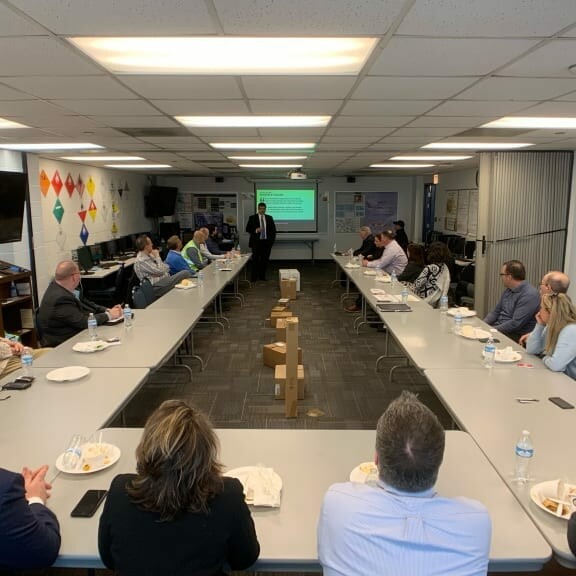 A group of people seated around a long rectangular conference table in a well-lit room are attentively listening to a person from K2 Solutions presenting information displayed on a screen. Some have laptops and notebooks in front of them, reminiscent of the focused discussions at the 2019 February Luncheon.