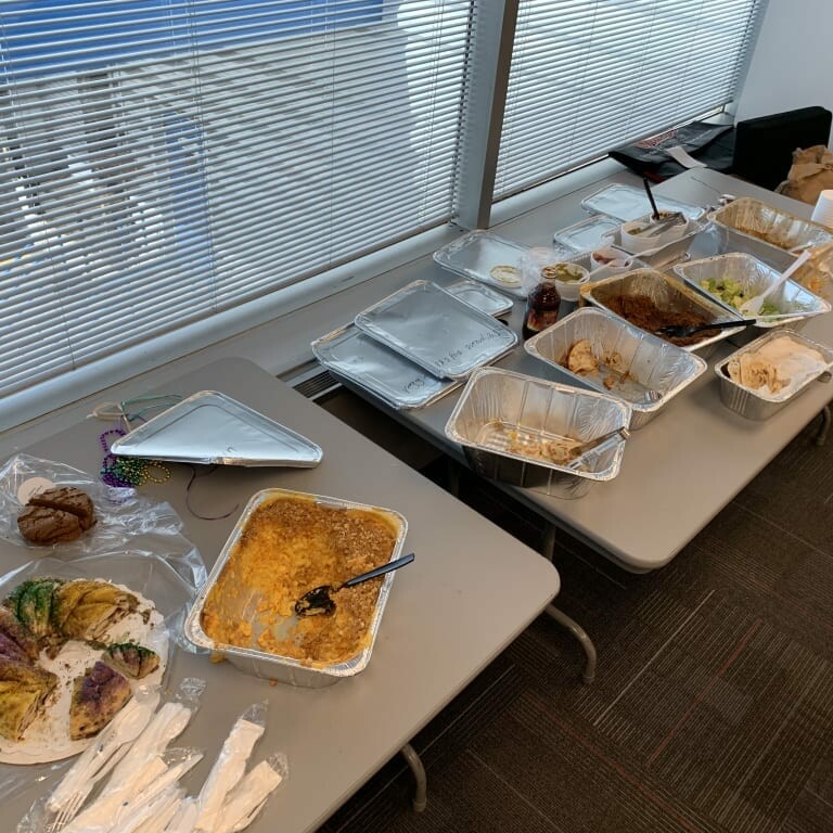 A long table in an office with remnants of the 2019 February Luncheon by K2 Solutions. Empty or partially empty trays, disposable cutlery, plates, napkins, and a few remaining food items such as pasta, bread slices, and cake are scattered across the surface. Blinds cover the windows behind.