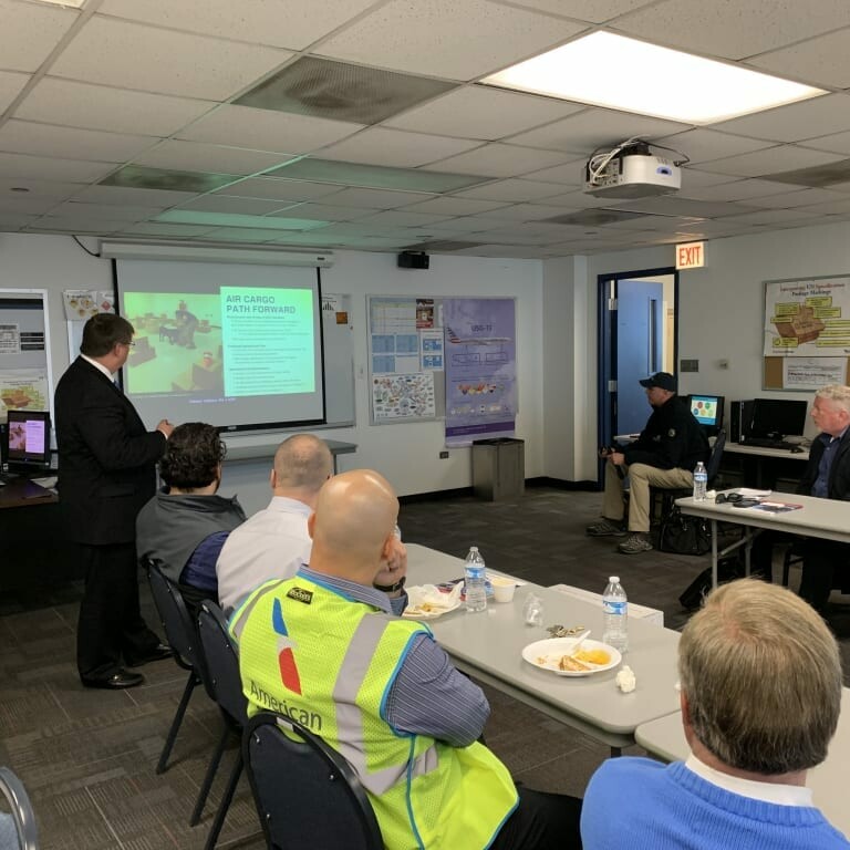 A group of people in a conference room are watching a presentation on a screen. The presenter, standing beside the slide titled "Air Cargo Plan Overview," points at it during the 2019 February Luncheon hosted by K2 Solutions. Some attendees are seated at tables with food and drinks in front of them.