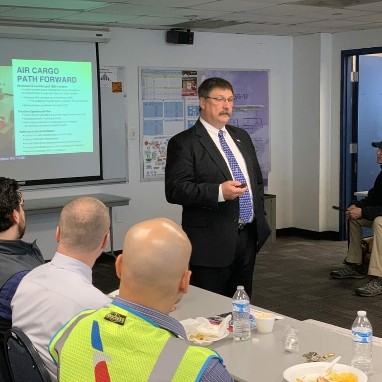 A man in a suit and tie speaks in front of a classroom setting. Behind him, a presentation slide titled "Air Cargo Path Forward" is displayed. Several people sit at tables, listening attentively during the 2019 February Luncheon featuring insights from K2 Solutions. The room has a mix of casual attire and uniforms visible.