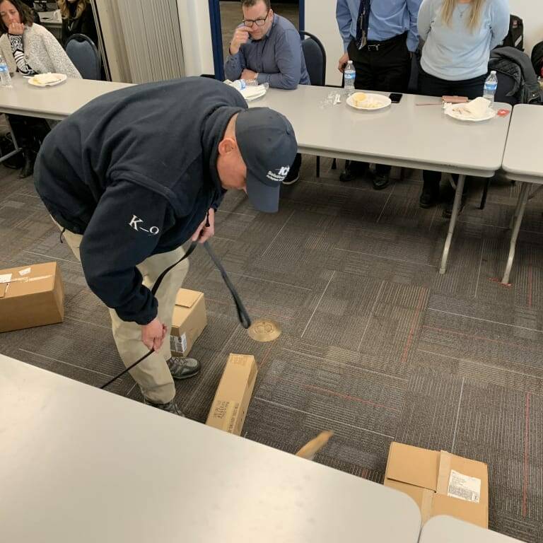 A man in a blue jacket and cap from K2 Solutions is using a metal detector to inspect a cardboard box on the floor. Several people are seated at tables around the room, observing him, suggesting this might be part of the 2019 February Luncheon or an indoor training session.