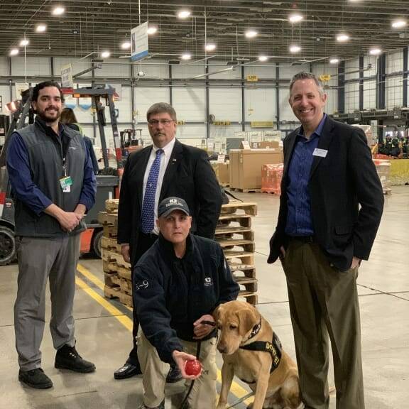 Four men are standing in a warehouse, smiling at the camera, while another man kneels, holding a red toy for a seated service dog. Pallets and shelves filled with boxes are visible in the background. This moment was captured during the 2019 February Luncheon hosted by K2 Solutions.