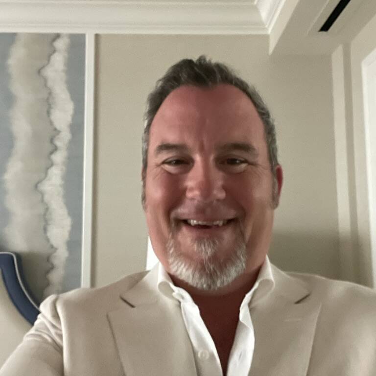 A middle-aged man with short gray hair and a neatly trimmed goatee smiles at the camera. William Gerst, a dedicated board member, wears a light-colored suit jacket and a white dress shirt. The background shows part of a room with a piece of abstract wall art and minimal decor.