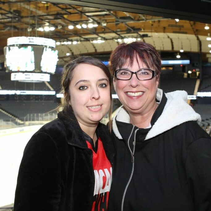 Two women smile for a photo inside an empty hockey arena during the 2018 March Networking Night. The woman on the left has long brown hair and wears a red jersey, while the woman on the right has short brown hair, glasses, and a black jacket. The ice rink and seating for the Chicago Wolves are visible in the background.