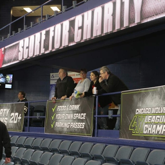 A group of people stand watching an event from an upper seating area in an arena. Banners advertise food, drinks, parking passes, and the Chicago Wolves' success as 4-time league champions. A digital sign above displays the message "SKATE FOR CHARITY" during the 2018 March Networking Night.