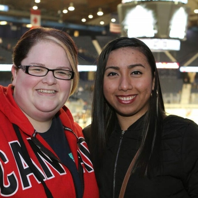 Two women are smiling at the camera in an indoor ice hockey arena during a Networking Night with the Chicago Wolves. The woman on the left is wearing a red Canada jacket, and the woman on the right is in a black jacket. The rink and spectators are visible in the background.