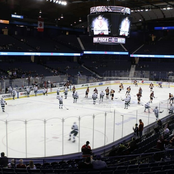 An ice hockey arena with players from two teams on the ice engaging in warm-up exercises. The stands have scattered spectators while the central scoreboard hangs above displaying information. The Canadian flag is visible among other advertisements around the rink during Chicago Wolves' Networking Night in 2018 March.