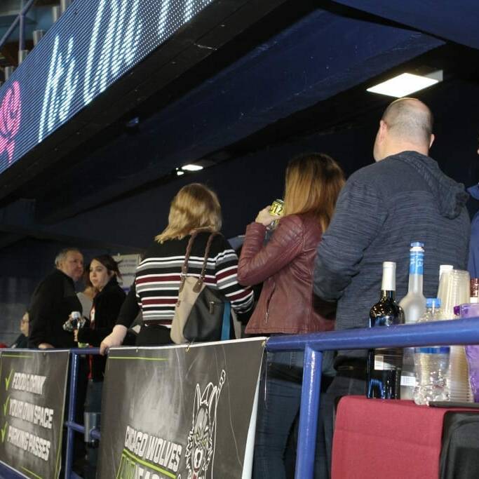 People are standing at a bar near a spectator area in an indoor arena during Networking Night. The bar counter has various drinks and bottles, with cups nearby. Overhead, a digital display reads "PARAMOUNT" with other text partially visible. The atmosphere appears casual, reminiscent of the Chicago Wolves' 2018 event.
