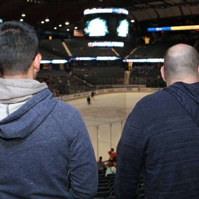 Two people with short hair, dressed in hooded sweatshirts, are seated in an arena watching a Chicago Wolves hockey game. The rink is brightly lit, and the scoreboard above displays the game details. The audience fills some seats in the background during 2018 March Networking Night.