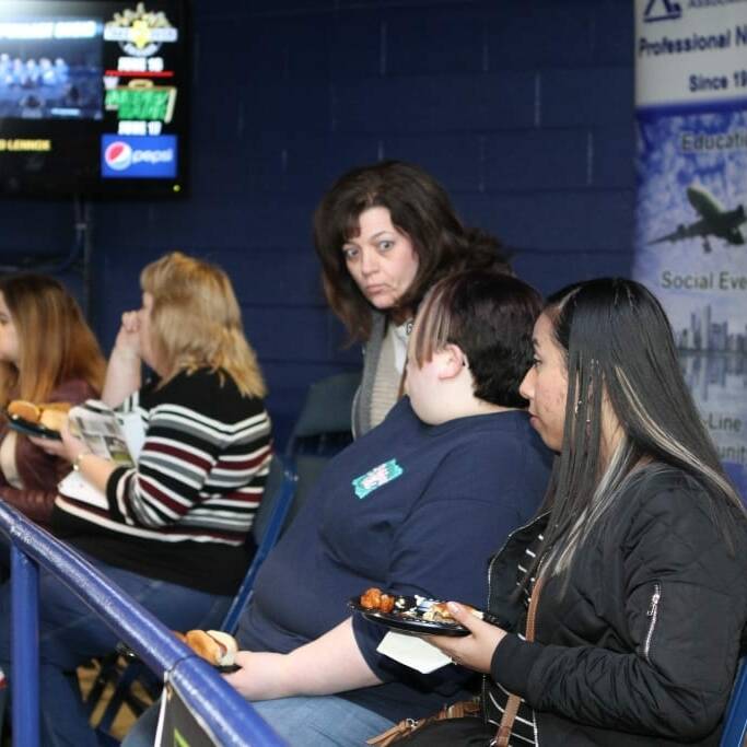 Several people are seated around a table at a networking event. Some are eating food from plates. Banners and a TV screen are in the background. One banner mentions "Professional Networking" and another features the word "Pepsi." The setting appears to be a casual 2018 March Networking Night hosted by the Chicago Wolves.